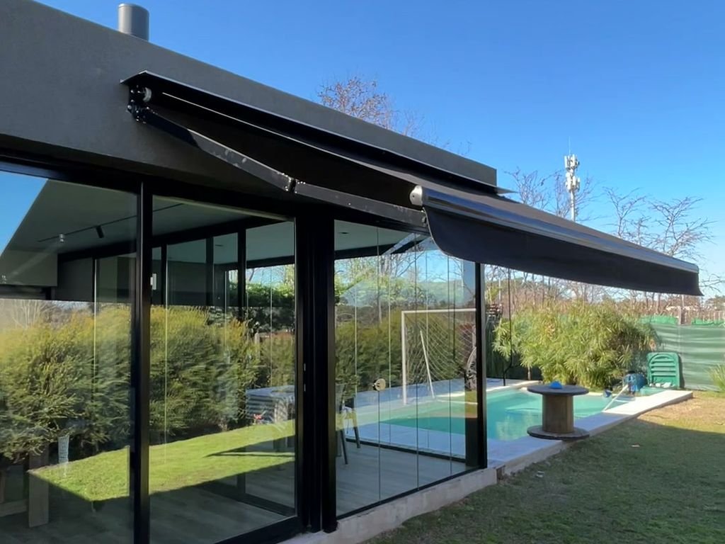 Toldo con brazo invisible negro desplegado sobre el ventanal de una casa con jardín y pileta
